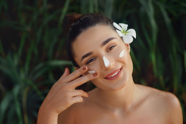 Concepto de belleza. Mujer aplicando crema cosmética, la extiende sobre su rostro para hidratar su piel y sonriendo.