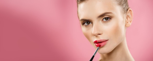 Concepto de belleza Mujer aplicando lápiz labial rojo con fondo de estudio rosa Hermosa chica hace maquillaje