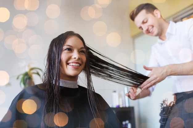 concepto de belleza, peinado y personas - mujer joven feliz y peluquera cortando puntas de cabello en el salón durante las luces de vacaciones