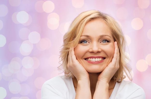 concepto de belleza, personas y cuidado de la piel - mujer sonriente de mediana edad con camisa blanca tocando la cara sobre fondo de luces de vacaciones rosas