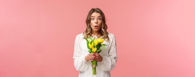 Concepto de belleza y primavera de vacaciones Retrato de niña rubia sorprendida y asombrada en vestido blanco sosteniendo tulipanes amarillos recibe flores siendo divertido y feliz de pie fondo rosa