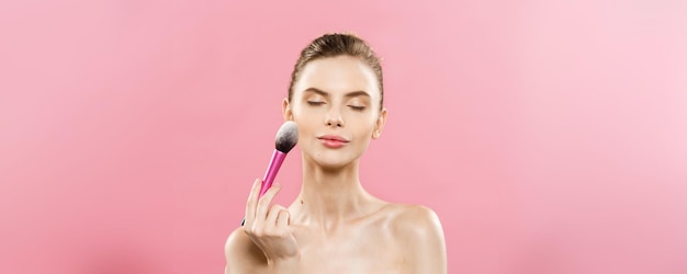 Concepto de belleza Primer plano Hermosa mujer caucásica aplicando maquillaje con brocha cosmética para polvo Piel perfecta aislada sobre fondo rosa y espacio de copia