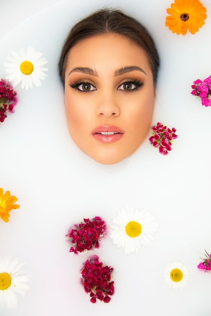 Concepto de belleza y spa auto indulgencia cuidado de la piel y el cuerpo. Hermosa mujer de piel clara y hermoso maquillaje en la bañera llena de leche con flores flotando.