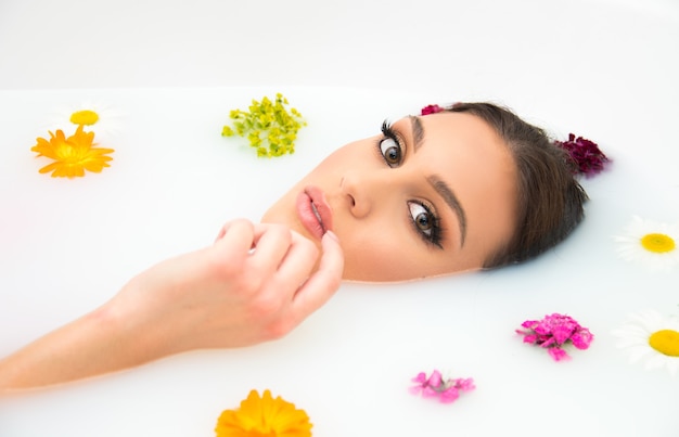 Concepto de belleza y spa auto indulgencia cuidado de la piel y el cuerpo. Hermosa mujer de piel clara y hermoso maquillaje en la bañera llena de leche con flores flotando.