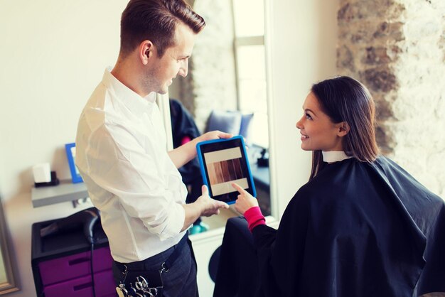 concepto de belleza, tecnología y personas - mujer joven feliz y estilista con computadora tablet pc eligiendo color de cabello en el salón