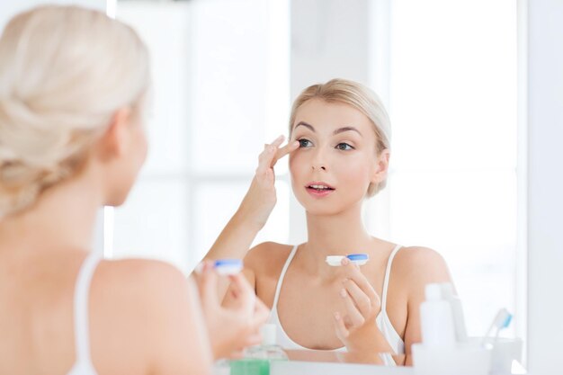 concepto de belleza, visión, vista, oftalmología y personas - mujer joven poniéndose lentes de contacto en el espejo del baño de casa