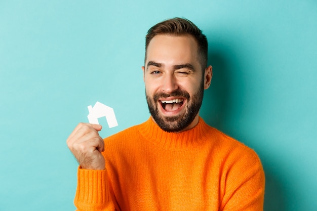 Concepto de bienes raíces. Feliz joven en busca de alquiler de casa, sosteniendo maket de papel de la casa y sonriendo, de pie sobre fondo azul.