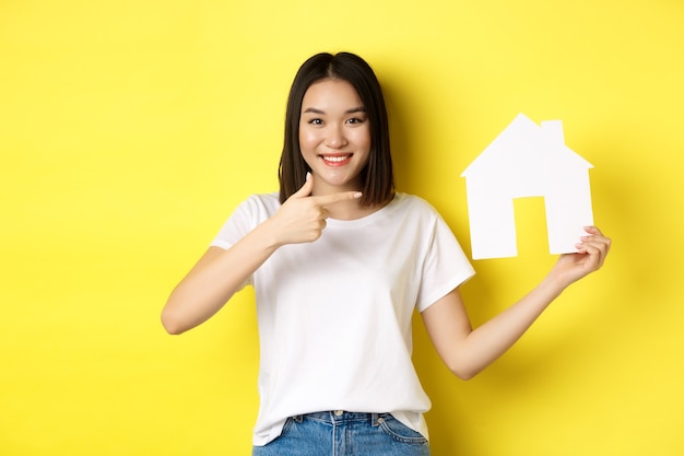 Concepto de bienes raíces y seguros. Alegre mujer asiática sonriendo, señalando el recorte de la casa de papel, recomienda el logotipo de la agencia, de pie sobre fondo amarillo.