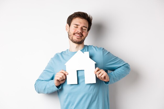 Concepto de bienes raíces y seguros. Soñador joven sonriendo con los ojos cerrados, mostrando el recorte de la casa de papel, deseando comprar una casa, de pie sobre fondo blanco.