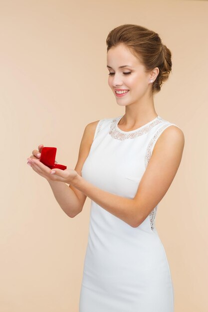 concepto de boda, amor, compromiso y felicidad - mujer sonriente con vestido blanco sosteniendo una caja de regalo roja con anillo de diamantes sobre fondo beige