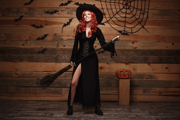 Foto concepto de la bruja de halloween - bruja del pelo rojo del feliz halloween que se sostiene que presenta con la escoba mágica sobre viejo fondo de madera del estudio.