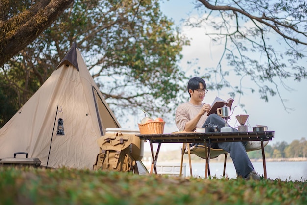 Concepto de camping Hombre acampando cerca del lago y sentado afuera de la tienda para relajarse leyendo un libro