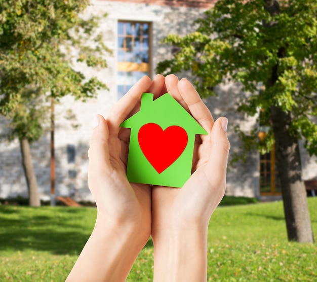 Foto concepto de caridad, bienes raíces y hogar familiar - imagen de primer plano de manos femeninas sosteniendo una casa de papel verde con corazón rojo