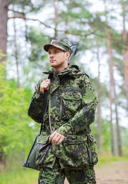 Concepto de caza, guerra, ejército y personas - joven soldado, guardabosques o cazador con pistola caminando en el bosque