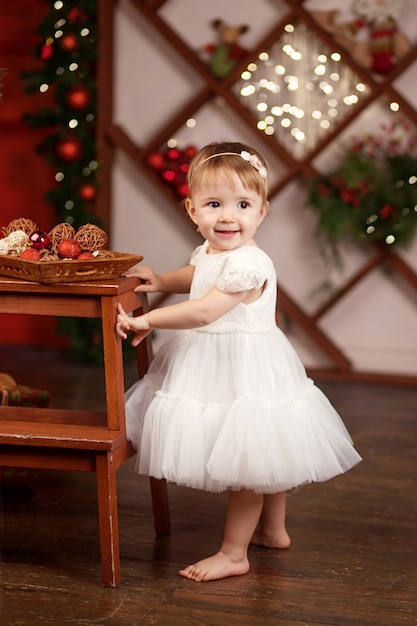 Concepto de celebración de año nuevo y Navidad. Niña bonita en vestido blanco jugando y siendo feliz con el árbol de Navidad y las luces. Vacaciones de invierno.