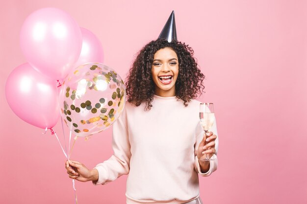 Concepto de celebración de cumpleaños - retrato de cerca de feliz alegre joven hermosa mujer afroamericana con camiseta rosa con coloridos globos de fiesta y champán
