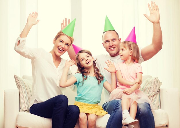 concepto de celebración, familia, vacaciones, niños y cumpleaños - familia feliz con dos niños con sombreros agitando las manos en casa