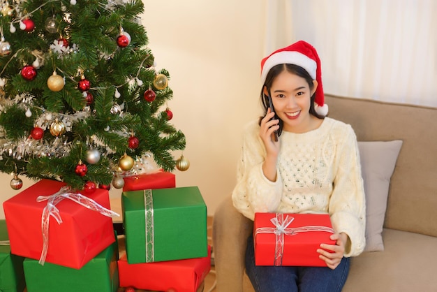 Concepto de celebración de navidad Mujer asiática joven sentada en la sala de estar y hablando por teléfono inteligente