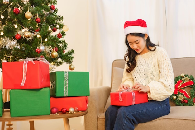 Concepto de celebración de navidad Mujer asiática joven sentada en el sofá y abriendo regalo de navidad