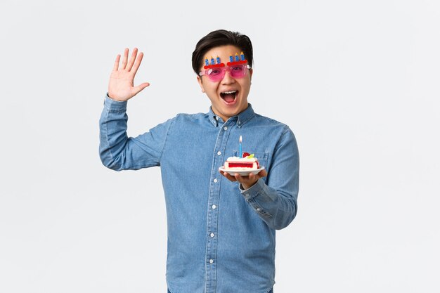 Concepto de celebración, vacaciones y estilo de vida. Feliz amigo sonriente en divertidas gafas de sol de fiesta trae pastel de cumpleaños con vela encendida, disfrutando del día b, divirtiéndose, de pie en la pared blanca