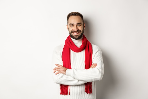 Concepto de celebración y vacaciones de Navidad. Hombre guapo con barba en suéter celebrando el año nuevo, con pañuelo rojo y sonriente, de pie sobre fondo blanco.