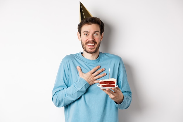 Concepto de celebración y vacaciones. Niño de cumpleaños sorprendido con sombrero de fiesta, sosteniendo pastel de cumpleaños y mirando agradecido, de pie sobre fondo blanco.