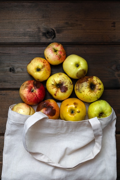 Concepto de cero residuos. Manzanas feas rojas y amarillas en el bolso blanco de la materia textil en la tabla de madera. manzanas esparcidas sobre la mesa. vista desde arriba.