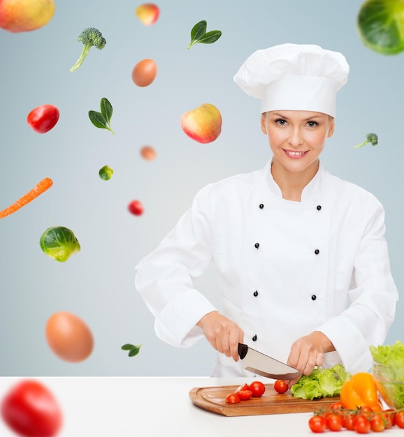 concepto de cocina y comida - chef sonriente, cocinera o panadera cortando verduras sobre fondo gris