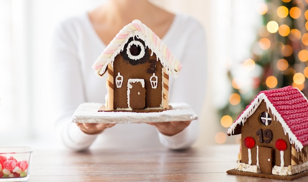 concepto de cocina, gente, navidad y horneado - cerca de una mujer feliz sosteniendo y mostrando una casa de pan de jengibre en casa