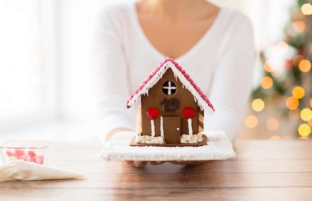 Foto concepto de cocina, gente, navidad y horneado - cerca de una mujer feliz sosteniendo y mostrando una casa de pan de jengibre en casa