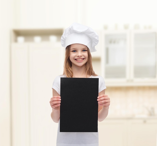 concepto de cocina, infancia, publicidad y personas - niña chef sonriente, cocinera o panadera con papel negro en blanco sobre el fondo de la cocina