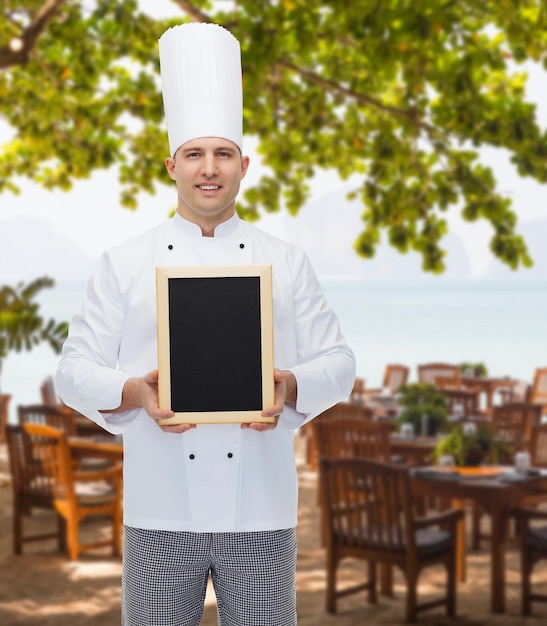 concepto de cocina, profesión, publicidad y personas - chef feliz mostrando y sosteniendo un tablero de menú en blanco sobre el salón del restaurante en la playa