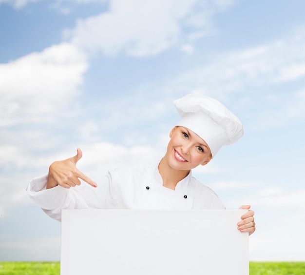 concepto de cocina, publicidad y personas: chef, cocinera o panadera sonriente señalando con el dedo a una pizarra blanca sobre el cielo azul y el fondo de la hierba