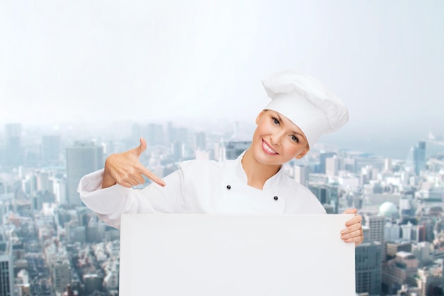 concepto de cocina, publicidad y personas - chef, cocinera o panadera sonriente señalando con el dedo a una pizarra blanca sobre el fondo de la ciudad