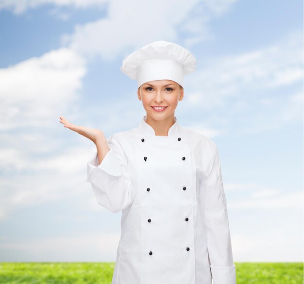 concepto de cocina, publicidad y personas - chef sonriente, cocinera o panadera sosteniendo algo en la palma de la mano sobre el cielo azul y el fondo de la hierba