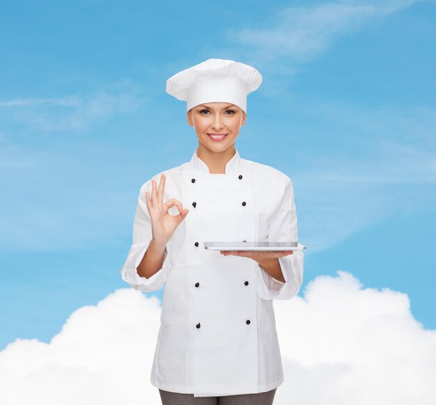 concepto de cocina, tecnología y personas: chef, cocinera o panadera sonriente con una computadora de tableta que muestra un gesto correcto sobre el cielo azul con fondo de nubes