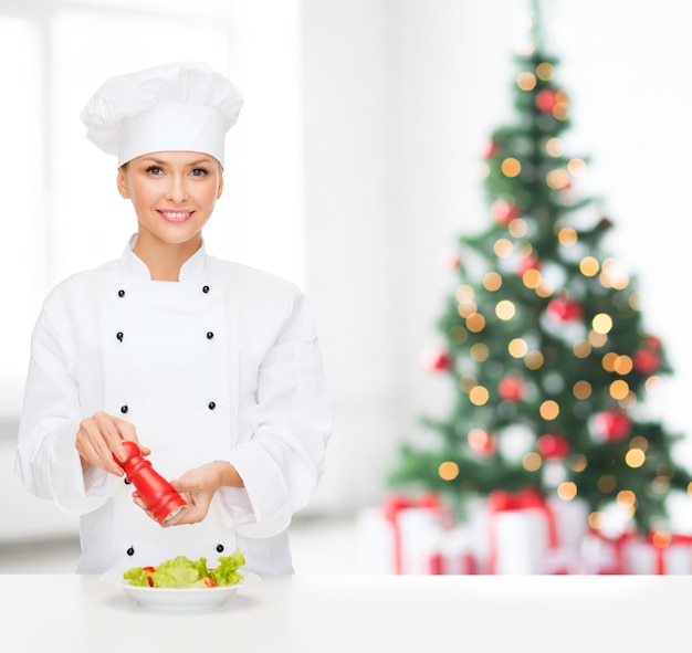 concepto de cocina, vacaciones, gente y comida - chef sonriente condimentando ensalada de verduras en la sala de estar con fondo de árbol de Navidad
