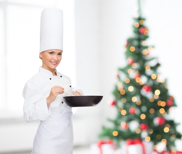 concepto de cocina, vacaciones, gente y comida - chef sonriente con sartén y cuchara mezclando comida sobre la sala de estar y el fondo del árbol de Navidad