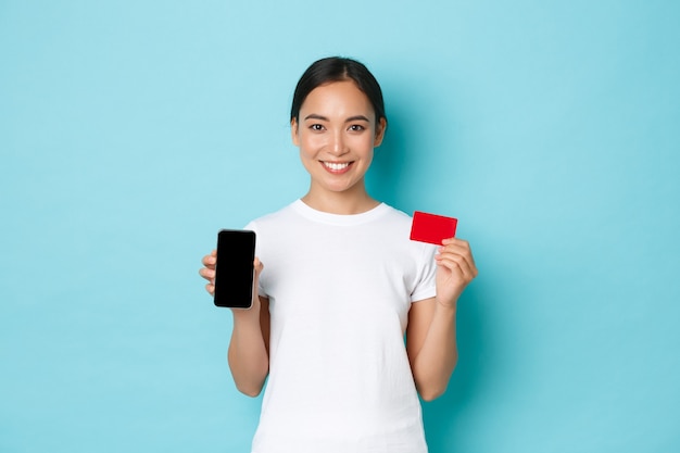 Concepto de comercio electrónico, compras y estilo de vida. Chica asiática alegre en camiseta blanca que muestra la tarjeta de crédito y la pantalla del teléfono inteligente, recomienda la aplicación para compras o transacciones en línea