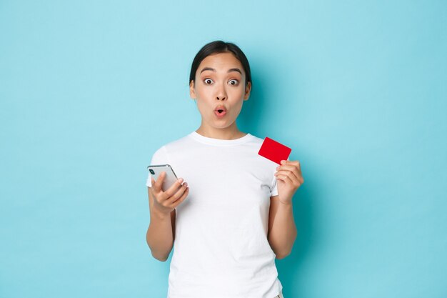 Concepto de comercio electrónico, compras y estilo de vida. Emocionada niña asiática sonriente se enteró de un descuento especial en línea, sosteniendo una tarjeta de crédito y un teléfono móvil, usando la aplicación para hacer un pedido, fondo azul claro.