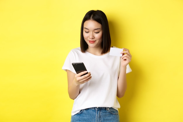Concepto de comercio electrónico y compras en línea. Hermosa mujer asiática pagando en internet, mirando la pantalla del teléfono inteligente y sosteniendo una tarjeta de crédito plástica, fondo amarillo