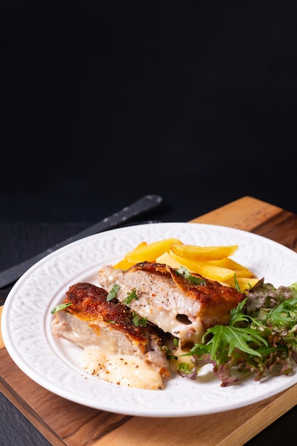 Concepto de comida Cordon bleu de ternera casera con ensalada y frito francés en placa de cerámica blanca sobre tabla de madera y fondo de piedra pizarra negra con espacio de copia