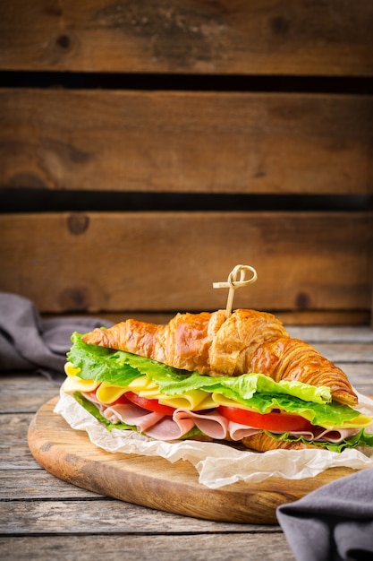 Concepto de comida, desayuno, mañana y almuerzo. Sándwich de croissant fresco con jamón, queso, lechuga y tomate en una mesa de madera