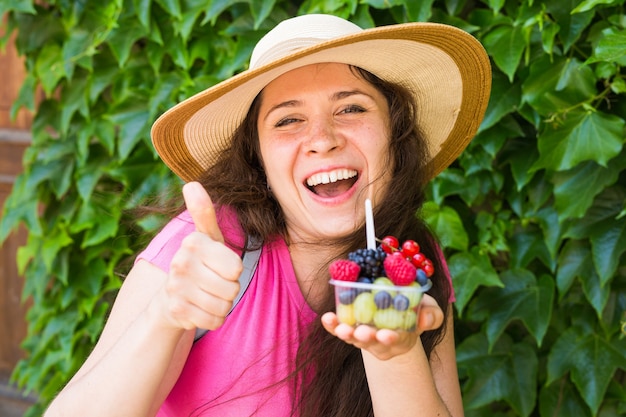 Foto concepto de comida ecológica y estilo de vida saludable - retrato de mujer sonriente sosteniendo bayas y mostrando el pulgar hacia arriba gesto.