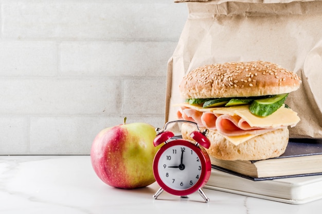 Concepto de comida escolar saludable, bolsa de papel con almuerzo, manzana, sándwich, libros y reloj despertador