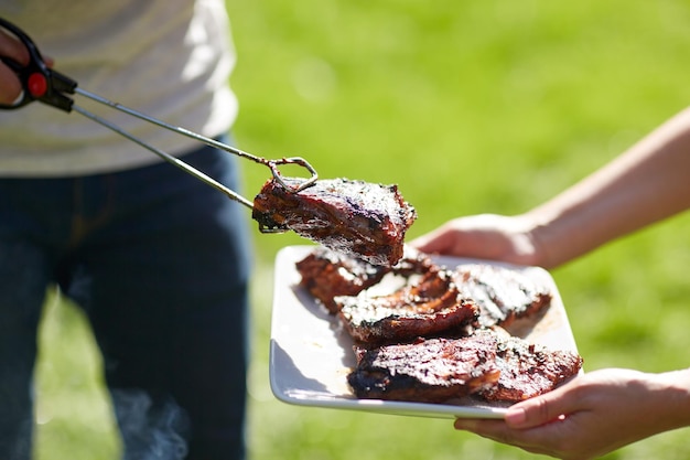 concepto de comida, gente, comida y cocina - hombre con pinzas poniendo carne de barbacoa en el plato en la fiesta de verano
