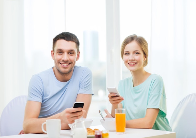 concepto de comida, hogar, pareja y tecnología - pareja sonriente con smartphones leyendo noticias y desayunando en casa