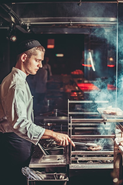 Concepto de comida joven chef guapo en uniforme blanco supervisa el grado de asado y convierte la carne