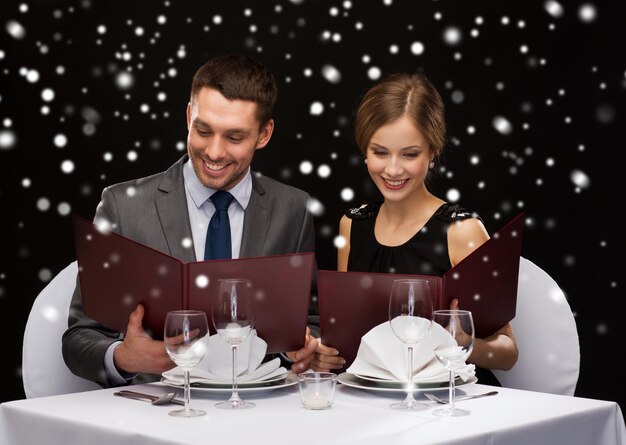 concepto de comida, navidad, vacaciones y personas - pareja sonriente con menús en el restaurante sobre fondo negro nevado