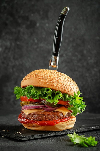 Concepto de comida rápida. Hamburguesa con cuchillo sobre tablero de pizarra sobre fondo oscuro.